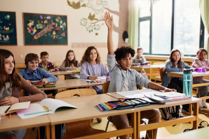 Student raising a hand in a bright elementary school classroom with classmates at desks