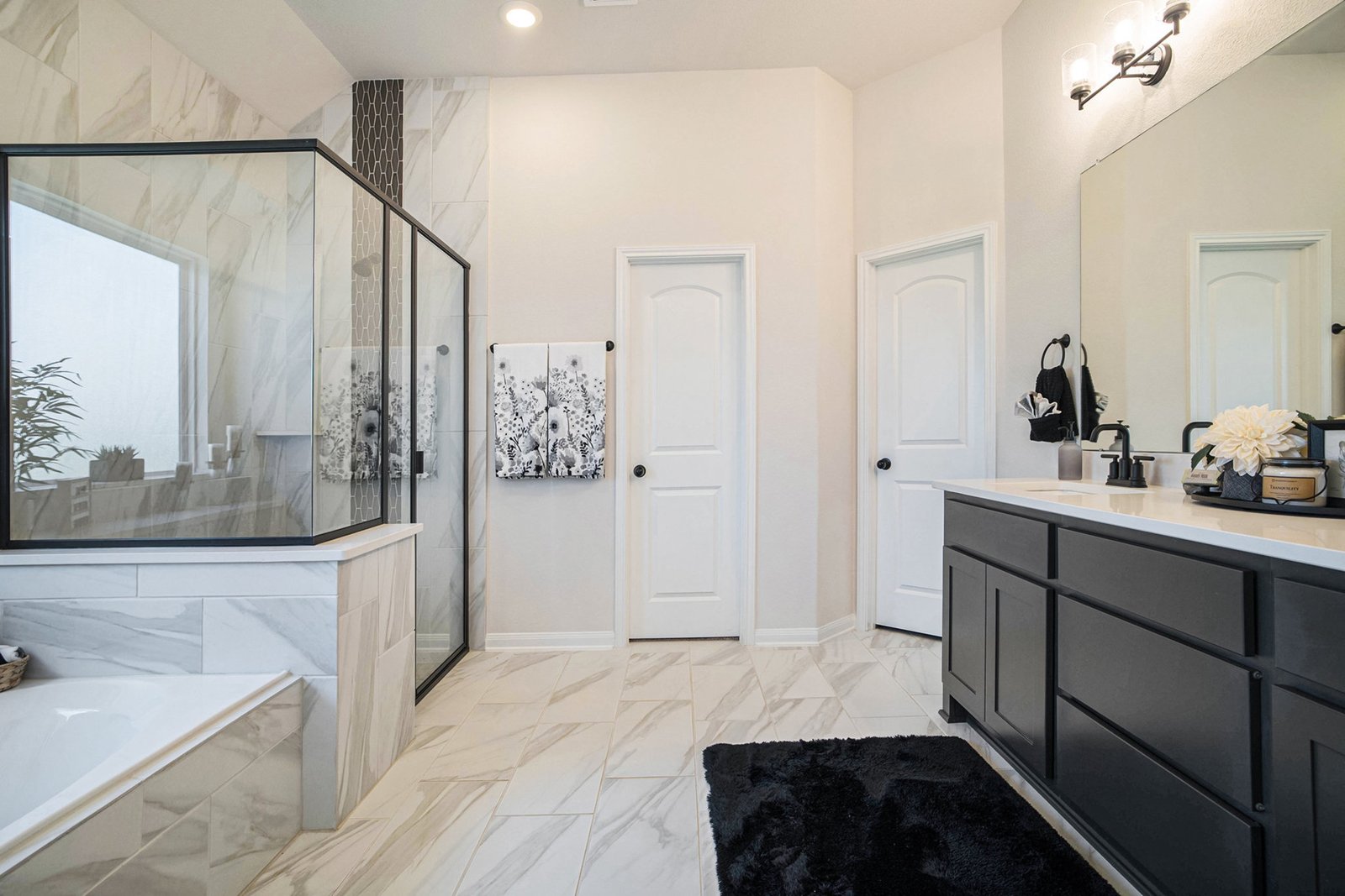 Primary bathroom with a marble-look tiled shower and corner tub, dark double vanity and a black bath mat