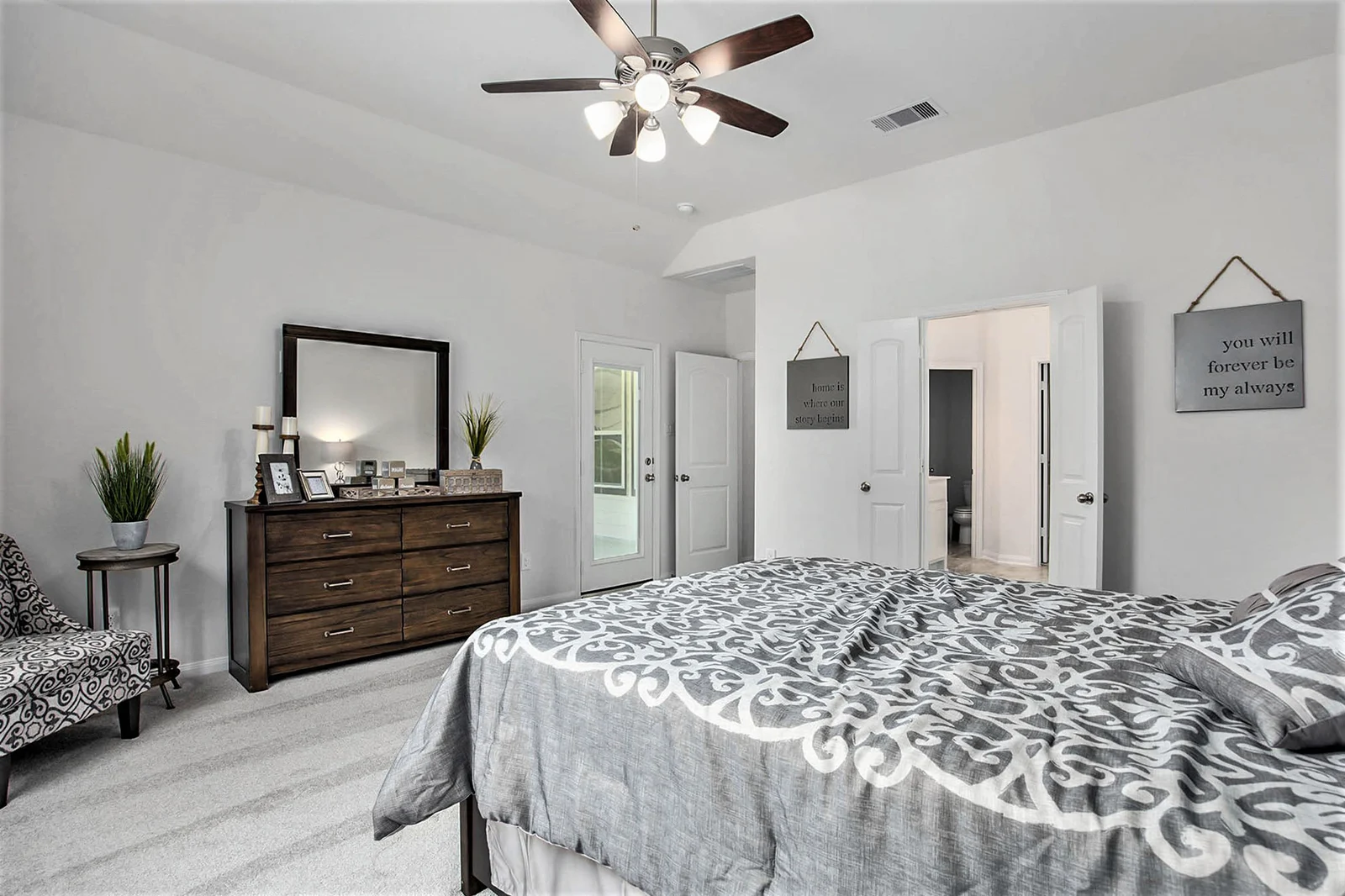 Kendall Homes model home primary bedroom with a dark wood dresser, ceiling fan and an en-suite bathroom beyond