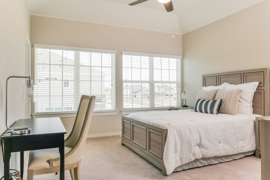 Carpeted bedroom with a grey timber bed frame, corner windows with blinds and a small desk