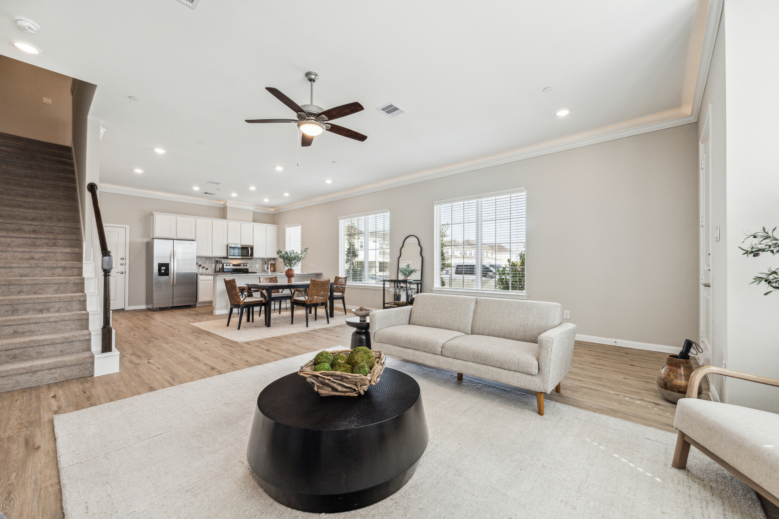 Open-plan living and dining room with a staircase, ceiling fan and a white kitchen beyond