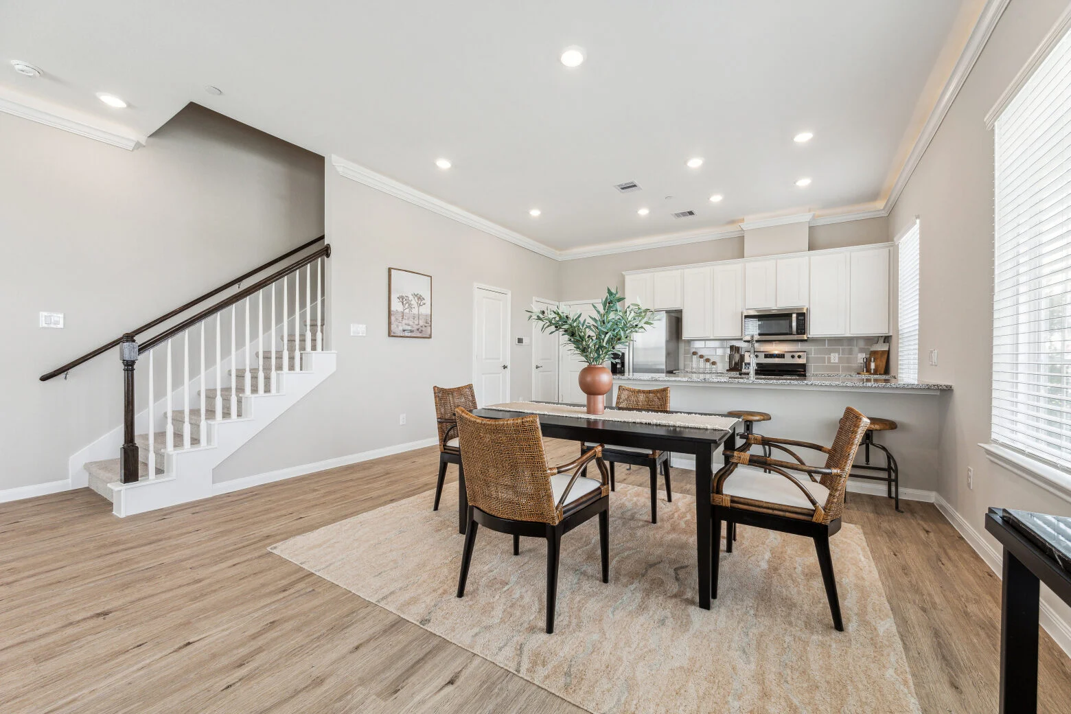 Dining area with a black table and rattan chairs beside a staircase, open to a kitchen with a breakfast bar