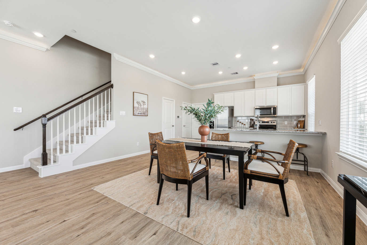 Dining area with a black table and rattan chairs beside a staircase, open to a kitchen with a breakfast bar
