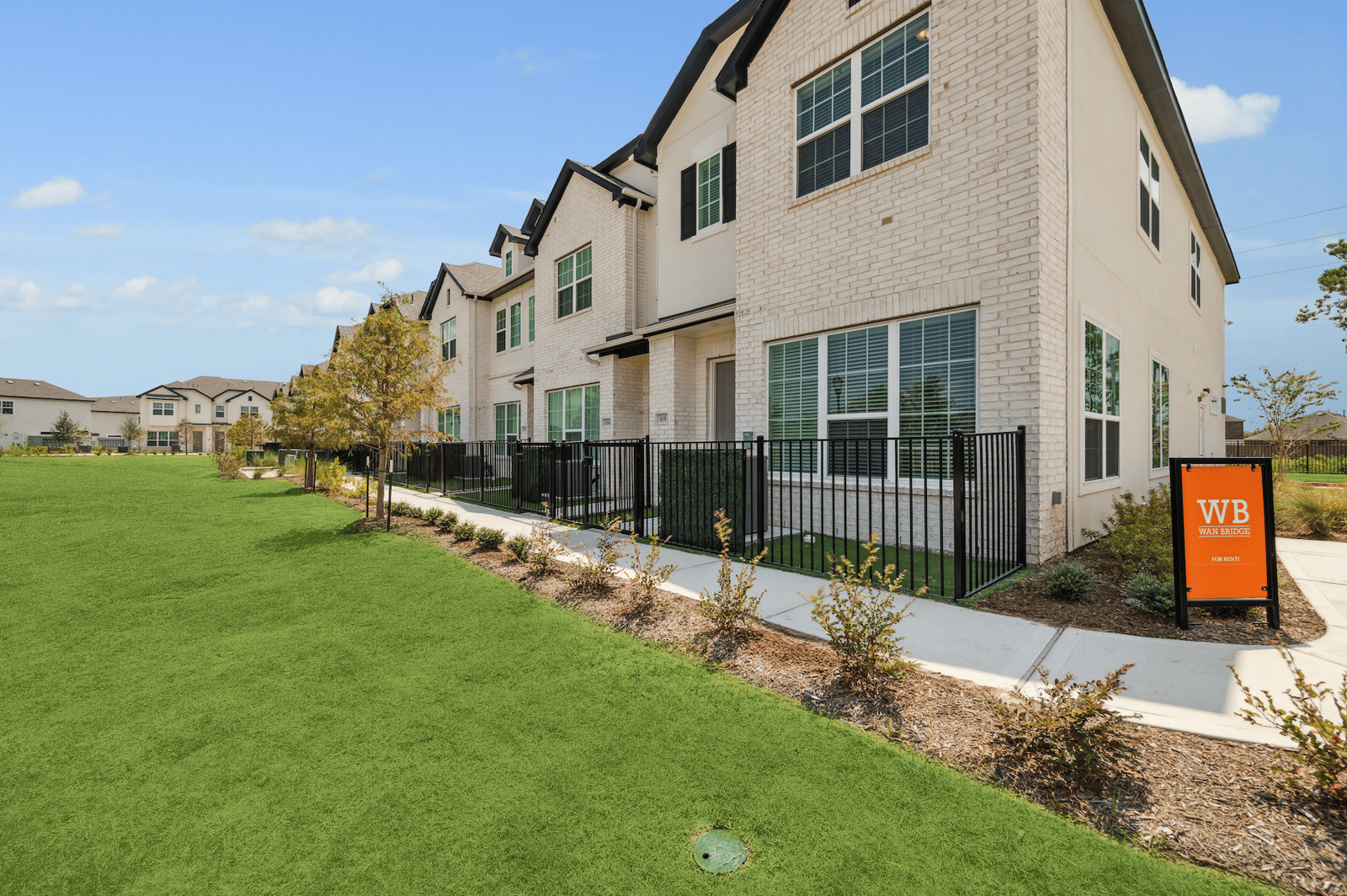 Row of pale-brick rental homes with fenced patios beside a lawn, with an orange WB Wan Bridge For Rent sign